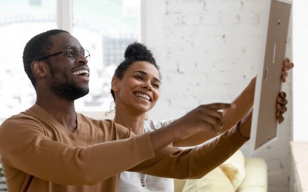 A couple smiling as they hang a framed picture on the wall of their home.